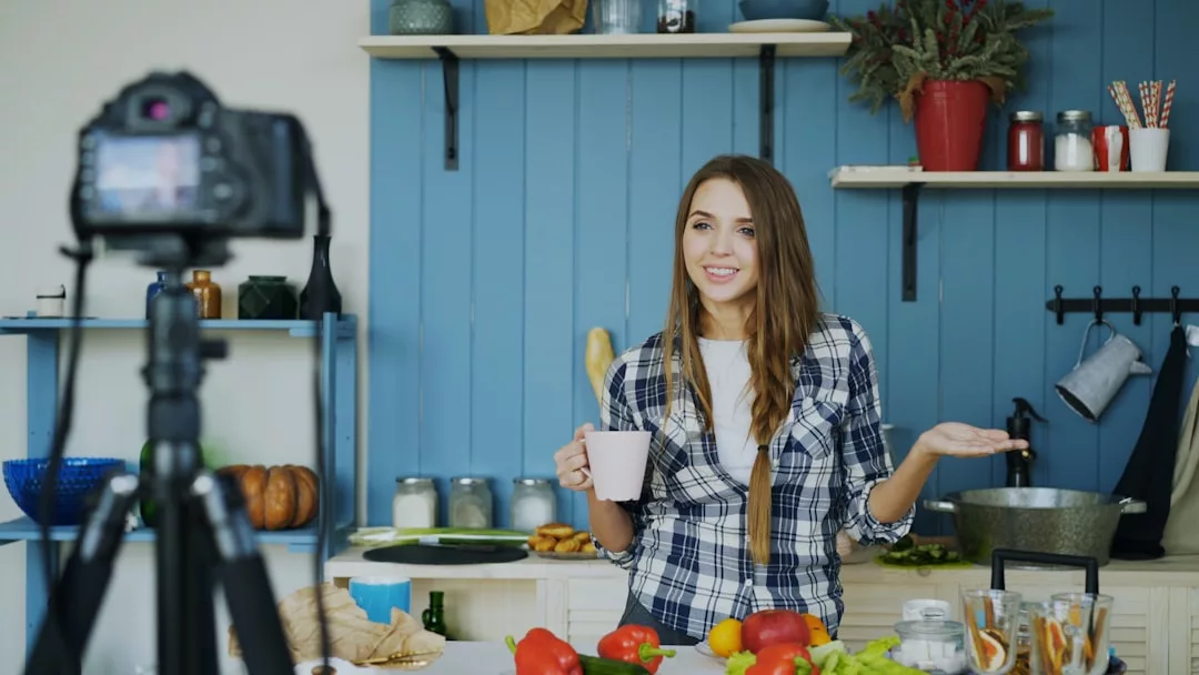 Eine Frau steht in einer Küche, hält einen Becher in der Hand und spricht in eine Kamera auf einem Stativ. Auf der Theke liegen Gemüse und Küchenutensilien, während sie über die neuesten Food-Trends berichtet.