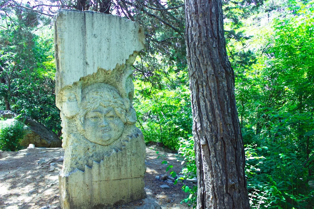 Eine verwitterte Steinskulptur mit einem geschnitzten menschlichen Gesicht steht im Freien zwischen Bäumen und Grün und erinnert an die zeitlose Eleganz der formellen Kunst in Deutschland.