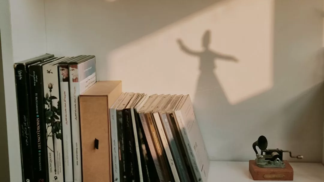 Ein Regal mit aufrecht stehenden Büchern und CDs, ein kleines antikes Grammophonmodell und ein Schatten an der Wand, der einer Person mit ausgestreckten Armen ähnelt, evozieren spirituelle Tiefe und die stille Essenz von Spiritualität.