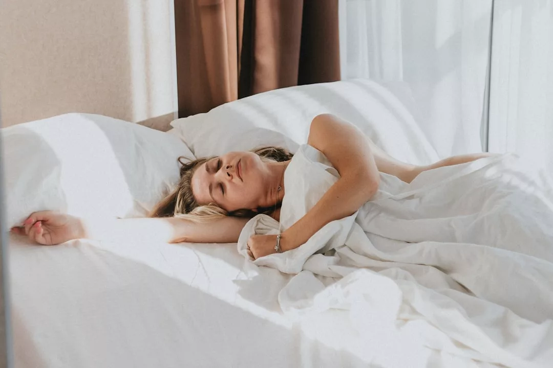Eine Person liegt im Bett unter weißen Laken, teilweise bedeckt vom Sonnenlicht, das durch ein Fenster fällt, und umarmt Tipps und Tricks zur Verbesserung der Schlafqualität.