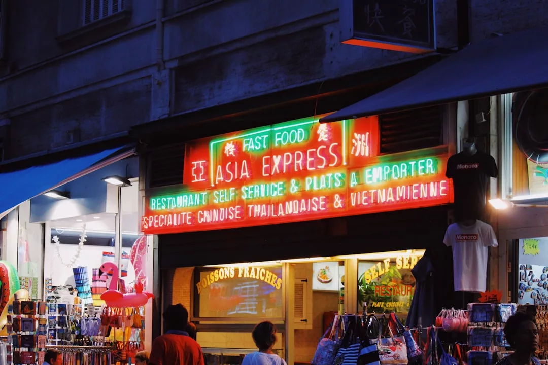 Neonleuchtschild für das Schnellrestaurant "Asia Express", das internationale Küche und Vielfalt mit chinesischen, thailändischen und vietnamesischen Gerichten anbietet, über einem Laden, in dem sich Menschen tummeln und Souvenirs verkauft werden.