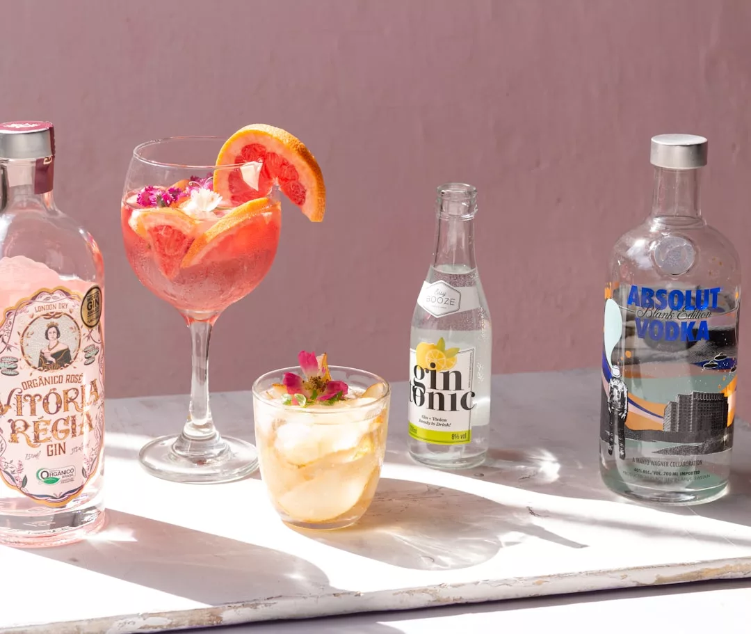 Eine Auswahl von Gin- und Wodkaflaschen mit zwei gemischten Cocktails - einer garniert mit Grapefruitscheiben und essbaren Blüten - auf einer weißen Fläche vor einem rosa Hintergrund.