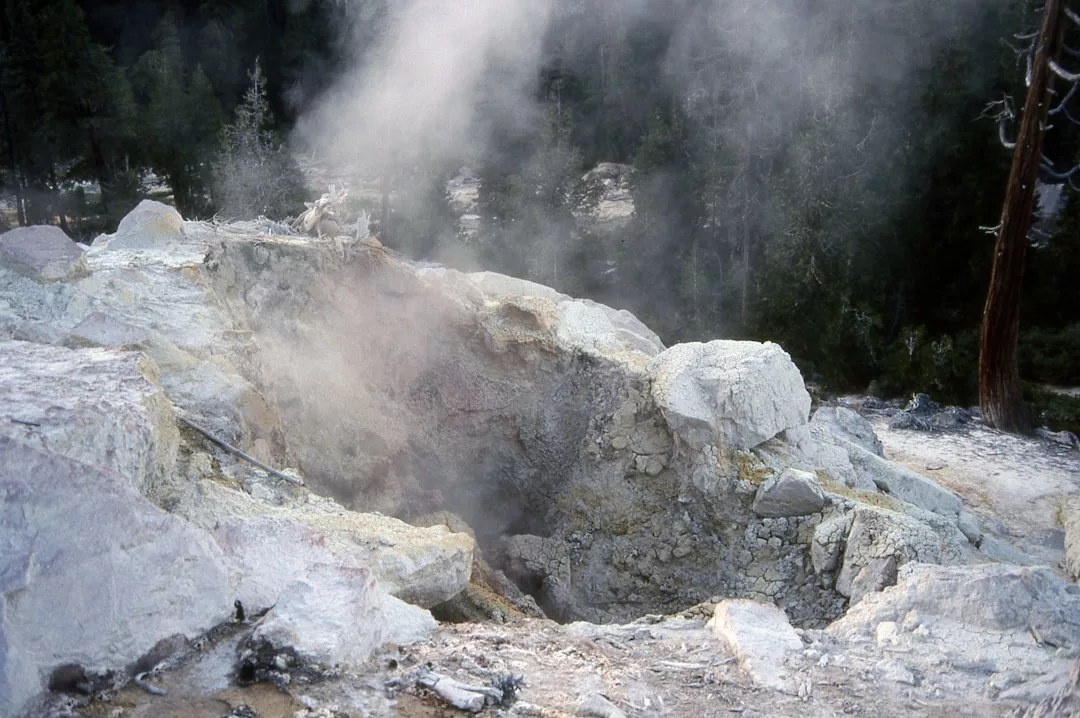 Eine felsige Fumarole stößt in einem bewaldeten Gebiet Dampf und Gase aus, mit sichtbaren Mineralablagerungen rund um die Öffnung - ein beeindruckendes natürliches Schauspiel der geothermischen Energie, ähnlich den Prinzipien der Erdwärmeheizung und ihrer Vorteile.