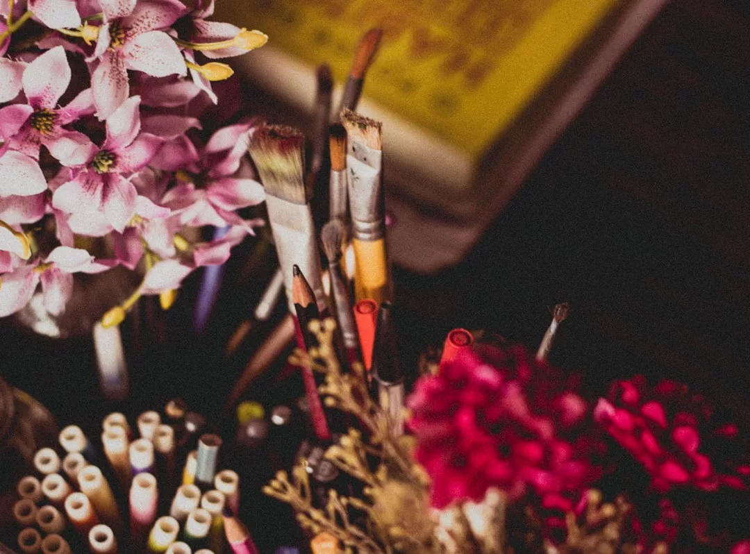 Nahaufnahme von verschiedenen Pinseln, Buntstiften und Markern in Behältern, umgeben von rosa und roten Kunstblumen - perfekt für die Kunsttherapie - mit einem verschwommenen Buch im Hintergrund.