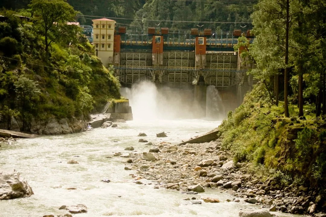 Ein Fluss fließt auf einen Staudamm zu, durch dessen Tore Wasser abgelassen wird, was die Vorteile der Wasserkraft zeigt, und das alles umgeben von Grün und bewaldeten Hügeln.