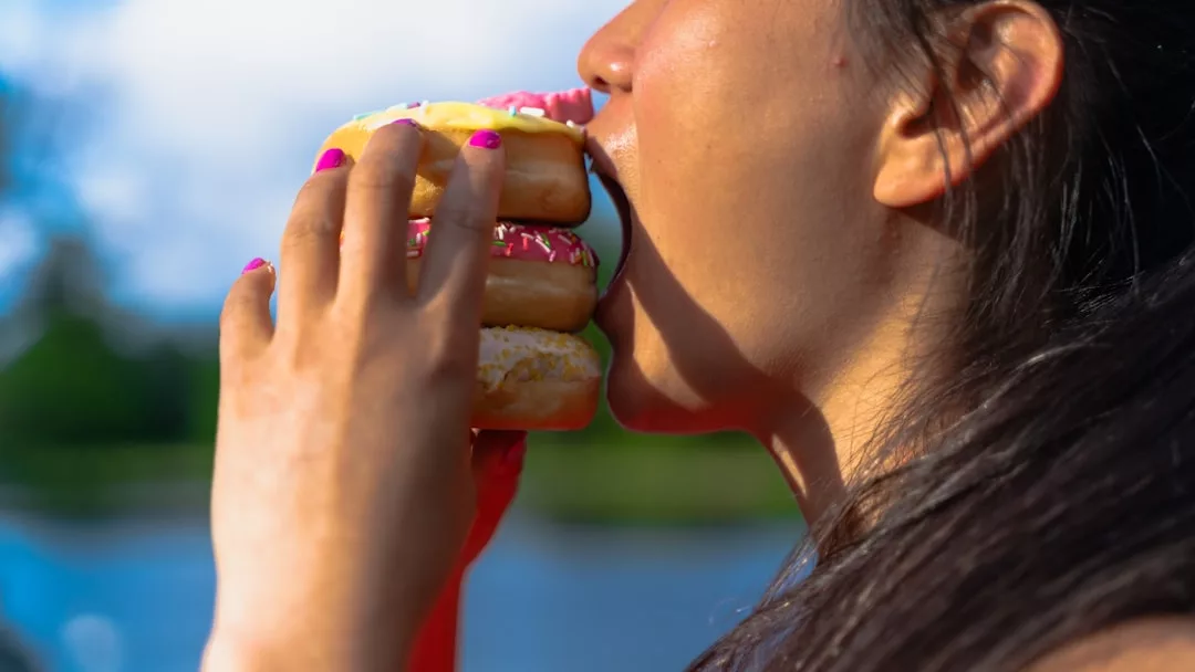 Eine Person mit langen Haaren und rosa Nagellack hält und beißt in einen Stapel von drei Donuts im Freien in der Nähe von Wasser. Sie gönnt sich einen Moment lang etwas, obwohl sie sich normalerweise auf gesunde Ernährung konzentriert, um ihr Immunsystem zu unterstützen.