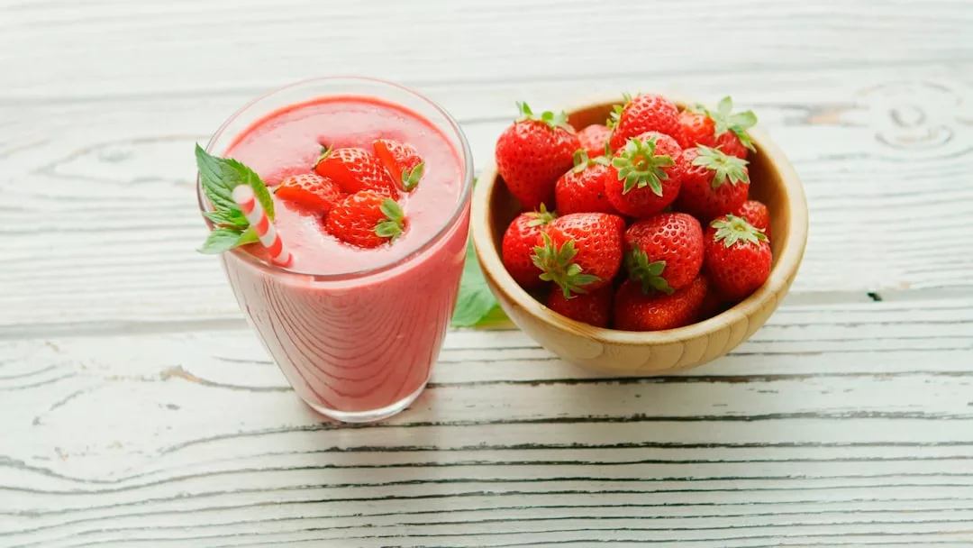Ein Glas Erdbeer-Smoothie garniert mit frischen Erdbeeren und Minze, daneben eine Holzschale mit ganzen Erdbeeren, auf einem weißen Holztisch - ein Genuss, der probiotische Ernährung und ihre Vorteile verbindet.