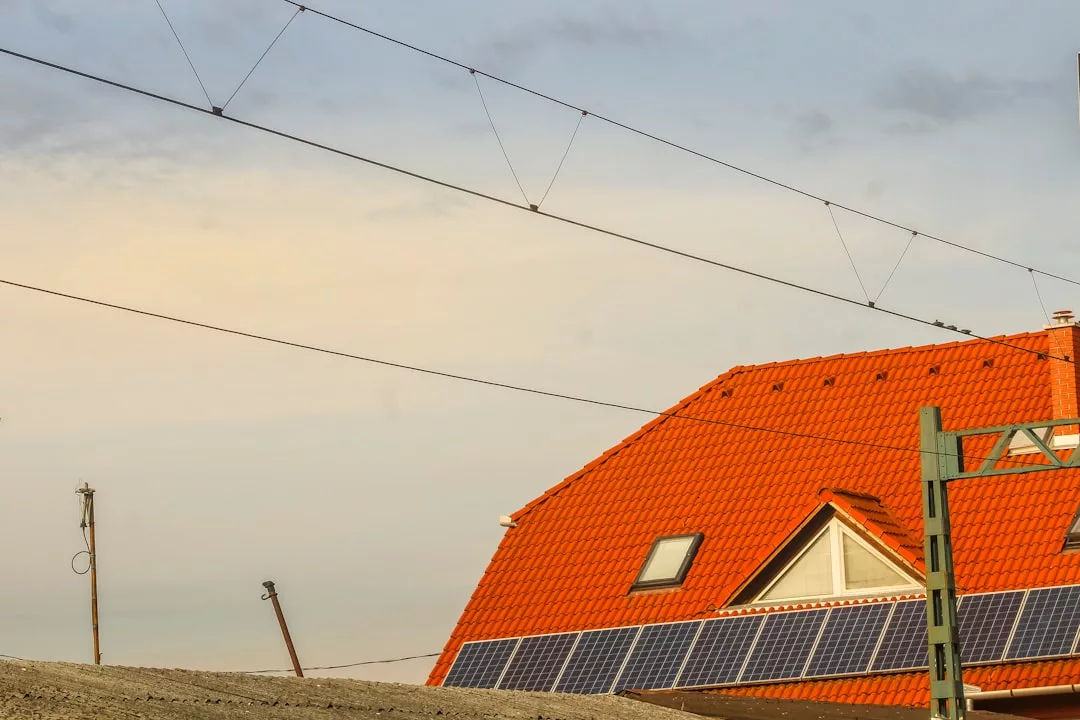 Rotes Ziegeldach mit Solarenergietechnik, durchkreuzt von Freileitungen vor teilweise bewölktem Himmel, zeigt die Vorteile von Solarenergie.