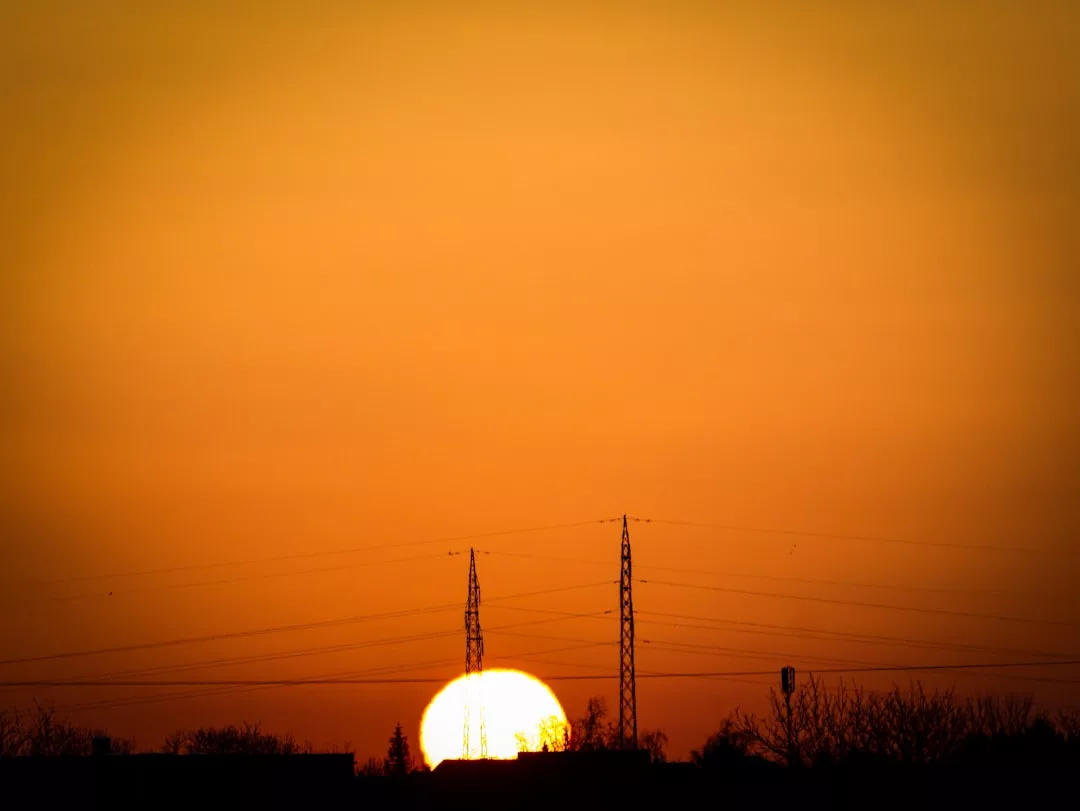 Die Sonne geht am Horizont hinter den Silhouetten von Stromleitungen und Bäumen unter und wirft ein orangefarbenes Licht auf den Himmel - eine schöne Erinnerung an die Vorteile und Potenziale der Solarthermie in der modernen Energiewirtschaft.