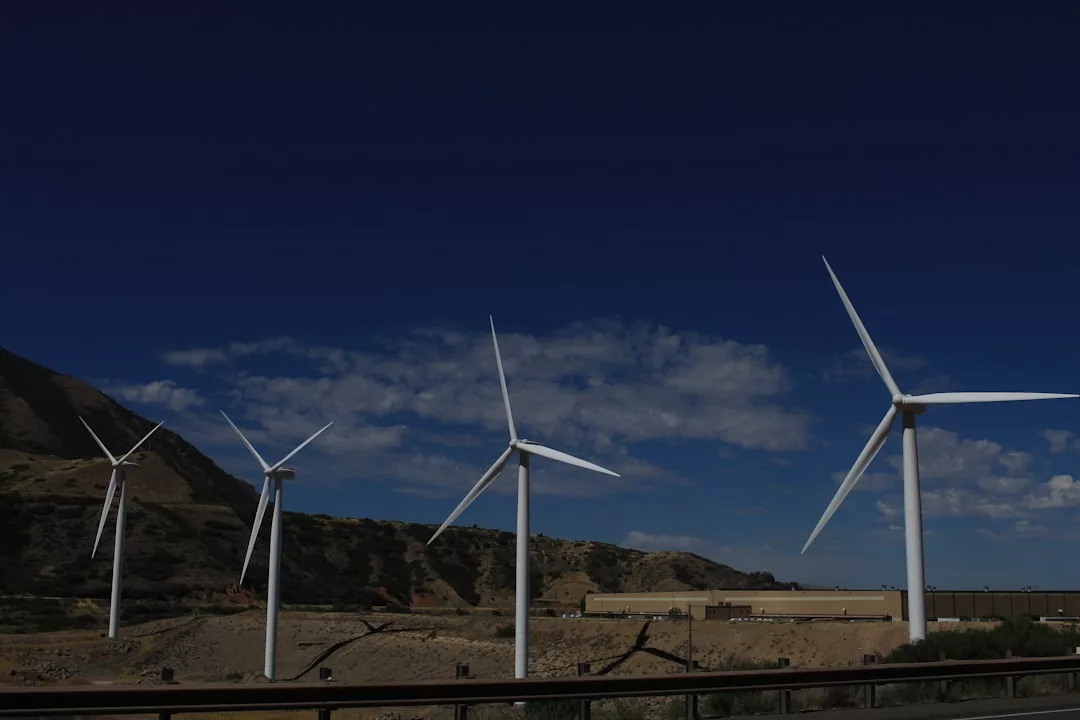 Vier Windturbinen stehen in einer Reihe in einer trockenen, hügeligen Landschaft unter einem klaren blauen Himmel und zeigen die Windkraftnutzung mit einem Gebäude und Bergen im Hintergrund.