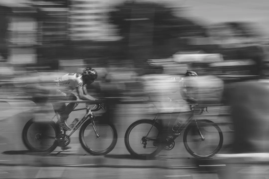 Zwei Radfahrer mit Helmen rasen auf einer unscharfen Outdoor-Strecke in Schwarz-Weiß, die die Geschwindigkeit und Bewegung des Fahrradfahrens einfängt und gleichzeitig seine Vorteile für Fitness und urbanes Leben hervorhebt.