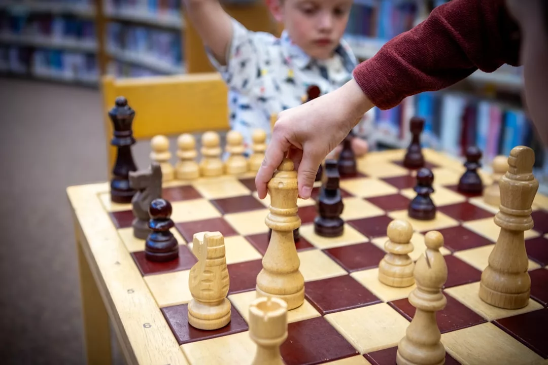 Ein Kind bewegt eine weiße Königsfigur auf einem hölzernen Schachbrett und zeigt strategisches Denken, während im Hintergrund ein anderes Kind und Bücherregale zu sehen sind.