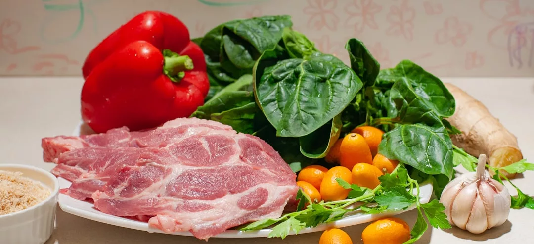 Ein Teller mit rohem Schweinefleisch, roter Paprika, Spinat, Kumquats, Ingwer, Knoblauch, frischen Kräutern und braunem Zucker präsentiert die Zutaten für eine proteinreiche Ernährung auf einer Theke.