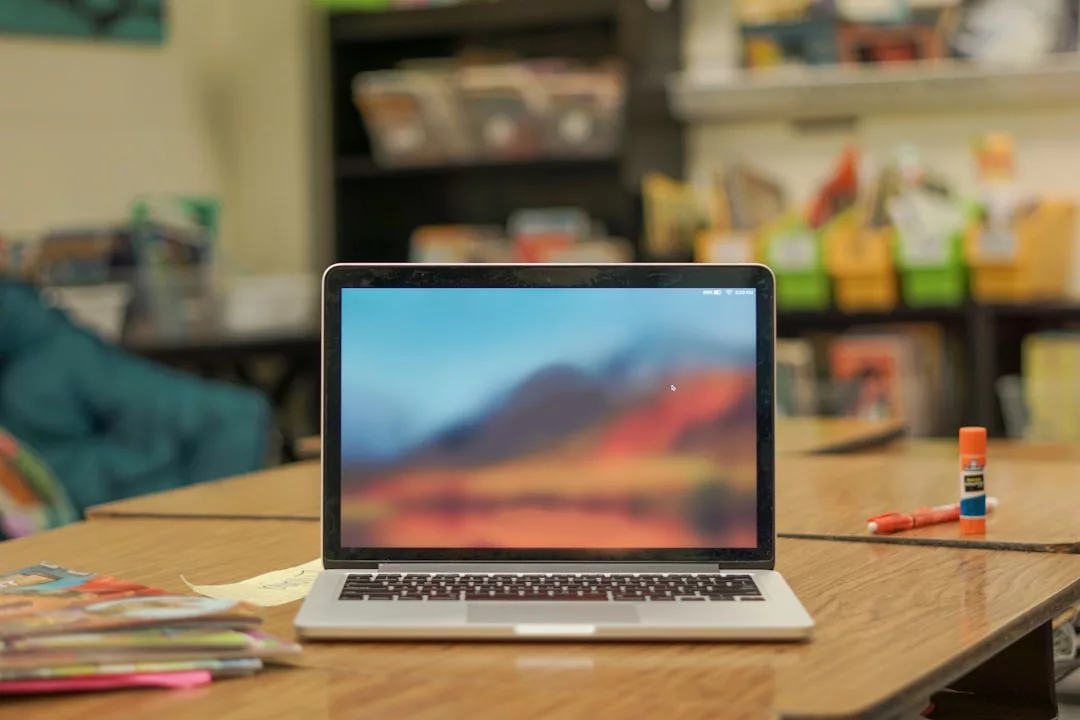 Ein Laptop mit unscharfem Bildschirm steht auf einem Tisch in einem Klassenzimmer, umgeben von Büchern, Papieren und Arbeitsmaterialien - zur Veranschaulichung der Vorteile von Fernstudium und Fernlernen in einer gut ausgestatteten Lernumgebung. Im Hintergrund sind Bücherregale und Mülleimer zu sehen.