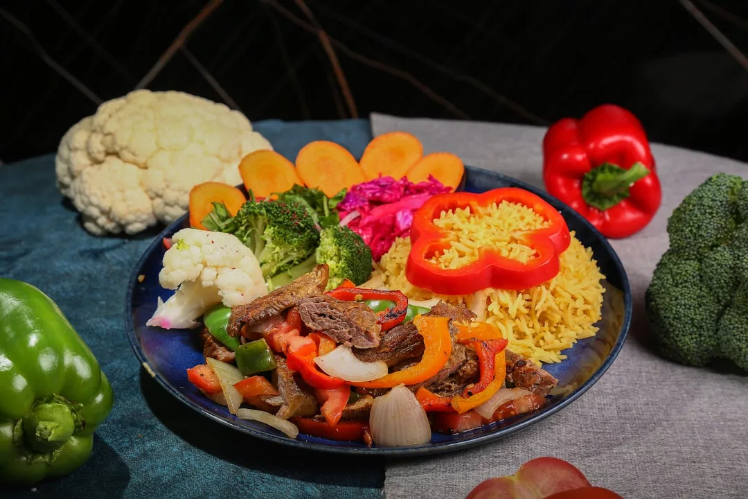 Ein Teller mit gelbem Reis, gebratenem Rindfleisch mit Paprika und Zwiebeln, gedünstetem Brokkoli, Blumenkohl, in Scheiben geschnittenen Karotten und einer Beilage aus lila Krautsalat - perfekt für Clean Eating und ein besseres Leben. Frisches Gemüse umgibt den Teller.