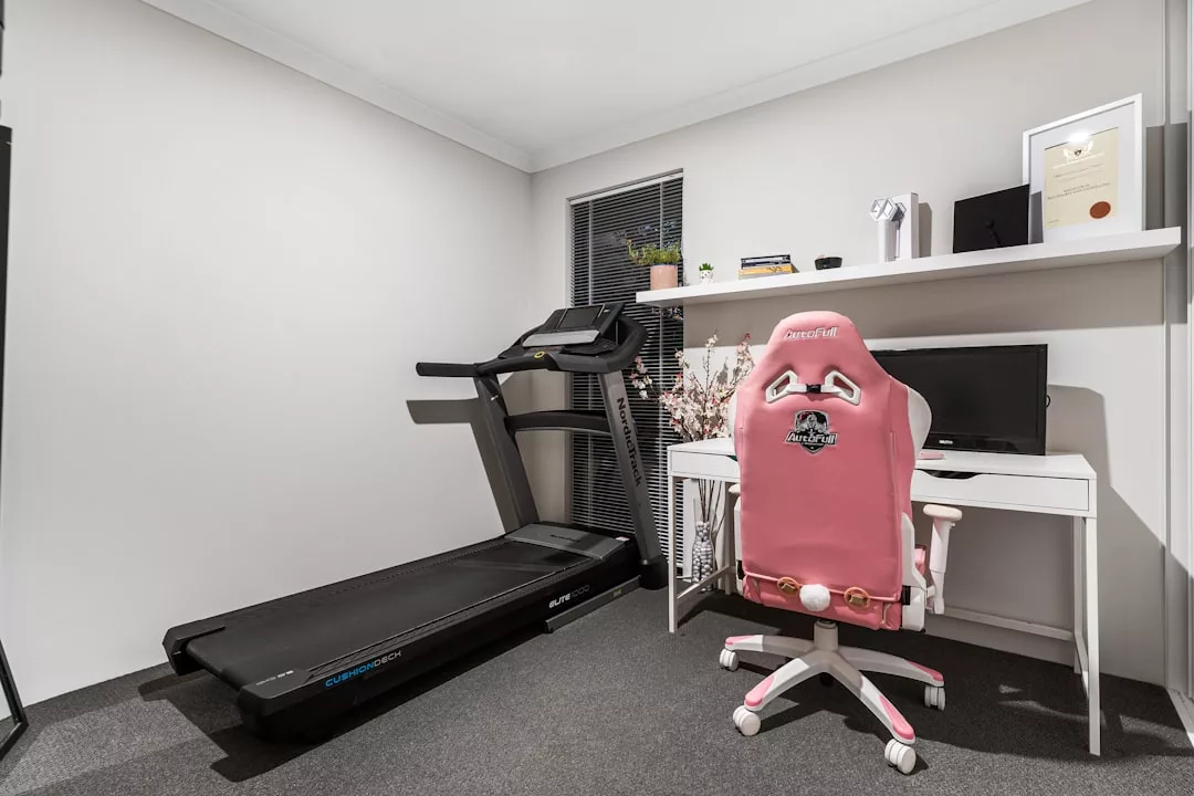 Ein Home-Office mit weißem Schreibtisch, Computer-Setup, rosa Gaming-Stuhl, Laufband, Regalen mit Dekoration und einem Fenster mit Jalousien - perfekt für effektives Training und Gesundheitsüberwachung dank integrierter Fitnesstracker-Technologie.
