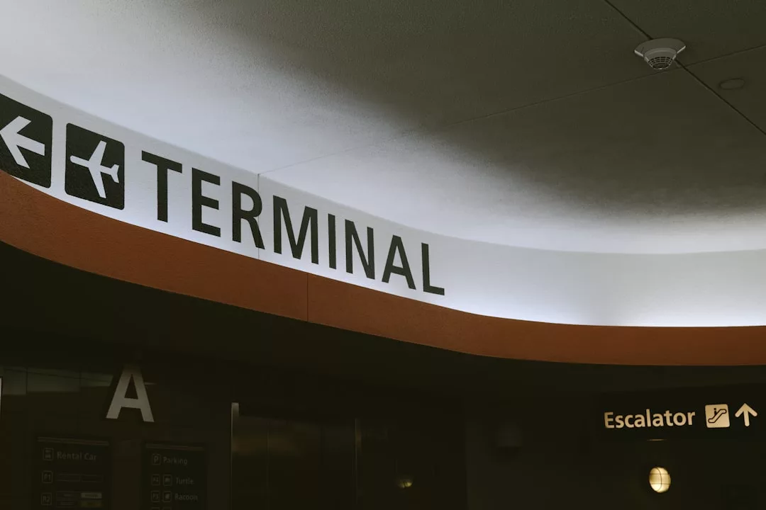 Ein Schild an einem Flughafen zeigt die Richtung zum Terminal mit einem Flugzeugsymbol und weist auf eine Rolltreppe hin, was auf die Vorteile von Langzeitreisen hinweist. Der Buchstabe "A" ist an einer Wand darunter zu sehen.