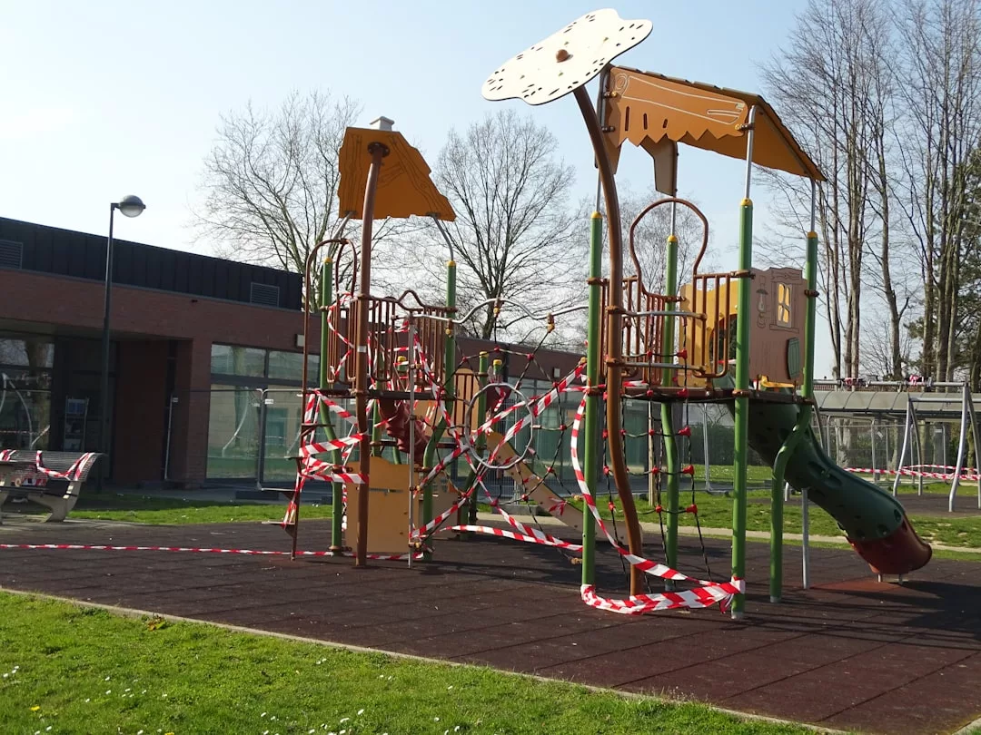 Spielplatzstruktur, umgeben von rotem und weißem Warnband, das anzeigt, dass der Spielplatz geschlossen oder gesperrt ist - was verdeutlicht, wie eingeschränkte regelmäßige Spielplatzbesuche die Vorteile eines Spielplatzes für die Kinderentwicklung beeinträchtigen können. Im Hintergrund sind Bäume und ein Gebäude zu sehen.