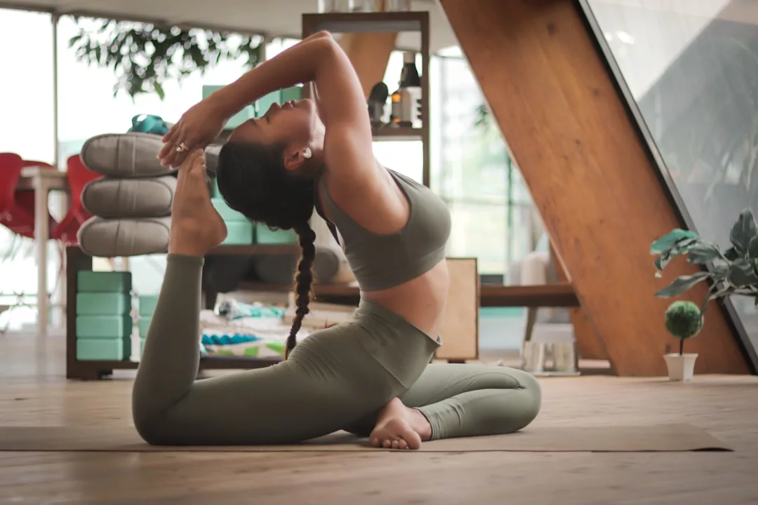 Eine Frau in Sportkleidung führt eine Yogapose in einem Innenraum aus, wobei sie ihren Fuß hinter dem Kopf hält, während sie auf einer Matte in einem gut beleuchteten Raum mit Holzfußboden sitzt, um die Vorteile von Yoga für die männliche Gesundheit und das allgemeine Wohlbefinden zu demonstrieren.