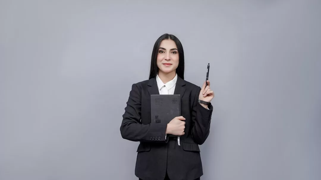 Eine Frau in einem schwarzen Anzug hält ein schwarzes Notizbuch in einem Arm und hebt mit der anderen Hand einen Stift, bereit, wichtige Fragen für ein Jobinterview zu beantworten, stehend vor einem schlichten grauen Hintergrund.