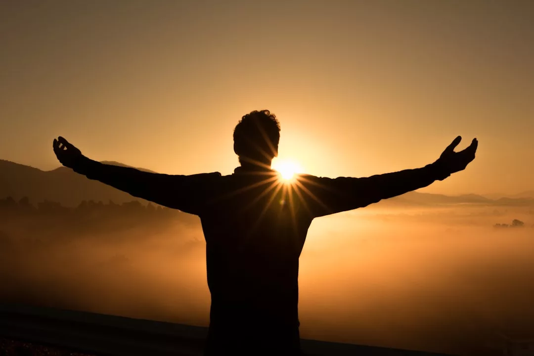 Silhouette einer Person, die bei Sonnenaufgang mit ausgestreckten Armen steht und die Wissenschaft des Glücks umarmt, während die Sonne hinter ihr über eine neblige Landschaft scheint.