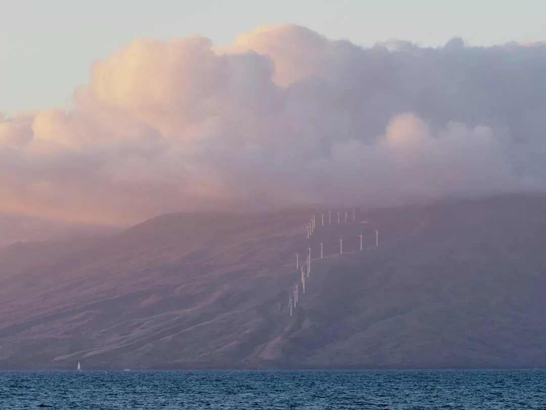 Eine Reihe von Windturbinen steht an einem Berghang unter einem wolkenverhangenen Himmel, mit dem Meer im Vordergrund - ein Symbol für die Zukunft der Energie und das Potenzial erneuerbarer Energien.