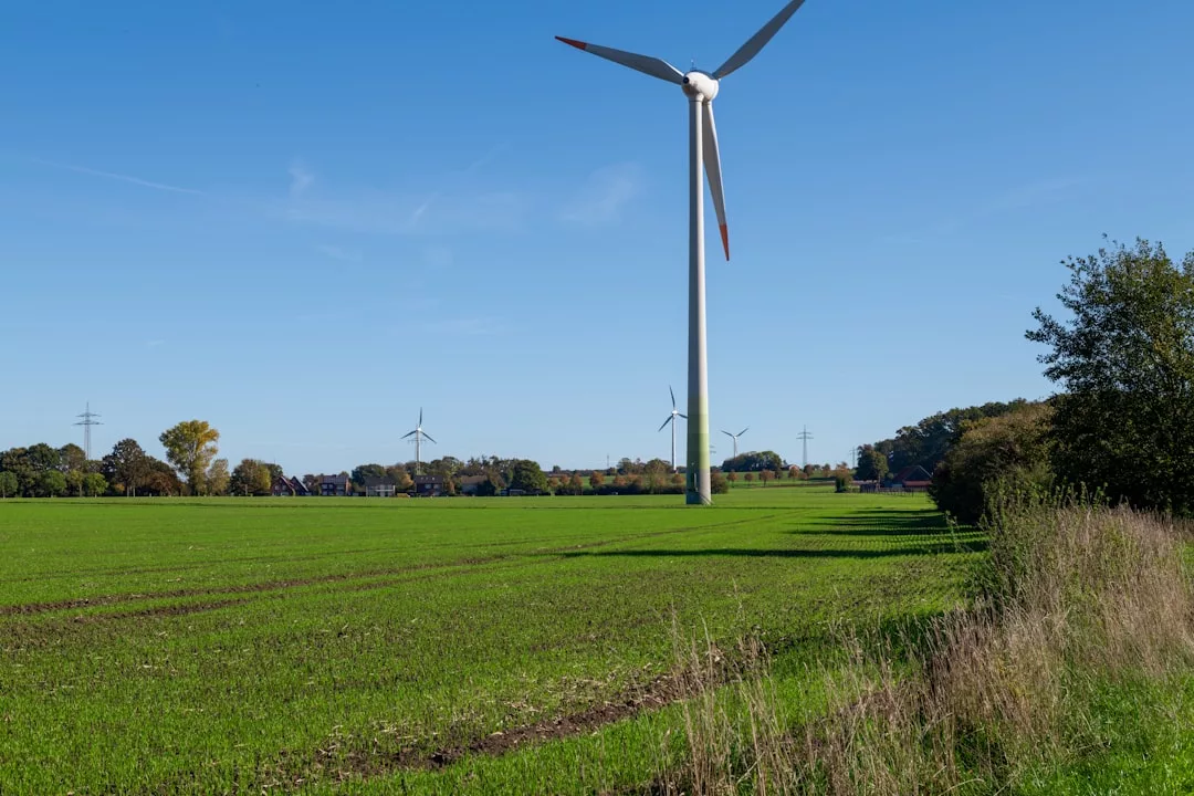 Eine Windkraftanlage steht auf einer grünen Wiese unter blauem Himmel und präsentiert innovative Windkrafttechnik. Im Hintergrund sind weitere Anlagen und Bäume zu sehen - eine Vision der nachhaltigen Zukunft.