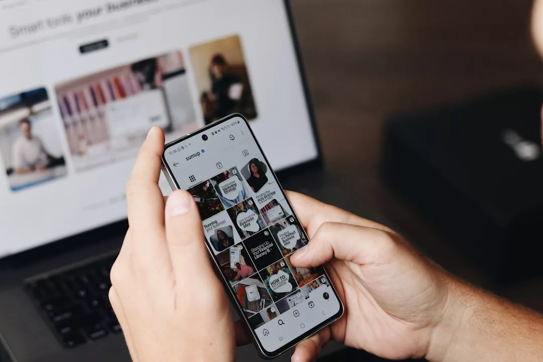 Eine Person scrollt auf einem Smartphone durch ein Instagram-Profil, während sie an einem Schreibtisch sitzt, an dem ein Laptop aufgeklappt ist und im Hintergrund die Website eines führenden Fintech-Unternehmens angezeigt wird.