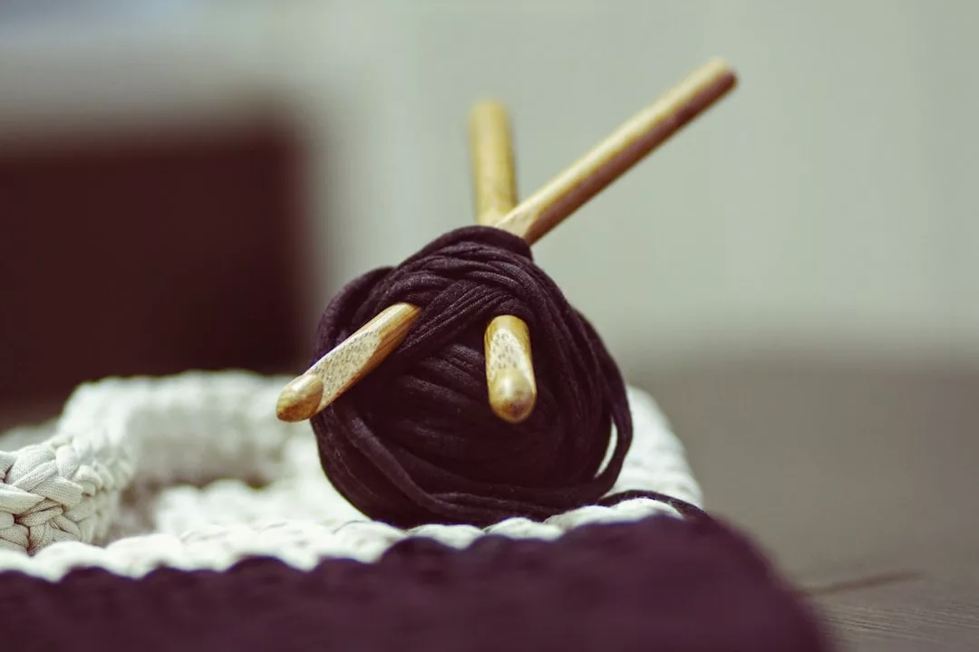 Eine Kugel aus dunklem Garn mit zwei Holz-Häkelnadeln steckt darin und liegt auf einem Stück gehäkeltem Stoff - perfekt für DIY-Projekte und kreative Ideen Zuhause.