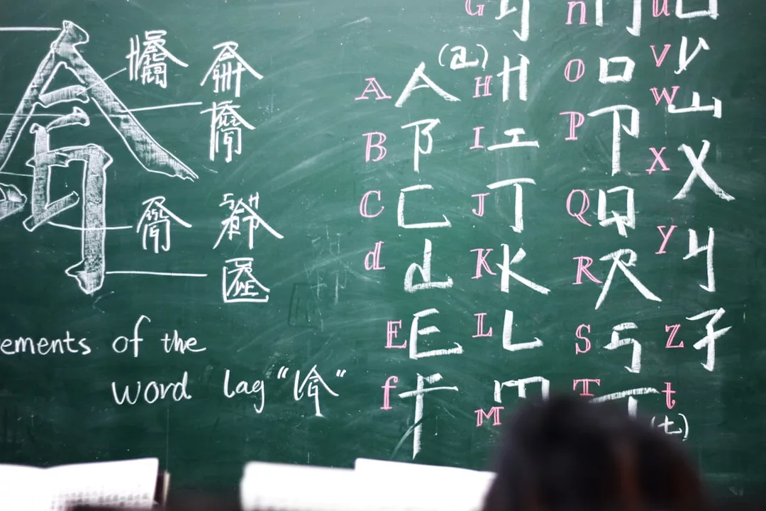 Auf einer Kreidetafel sind chinesische Schriftzeichen mit schematischen Bestandteilen und eine englische Alphabettafel mit den entsprechenden chinesischen Buchstaben abgebildet, so dass E-Learning-Elemente mit dem traditionellen Klassenzimmer verschmelzen; am unteren Rand erscheinen teilweise Schülerköpfe.