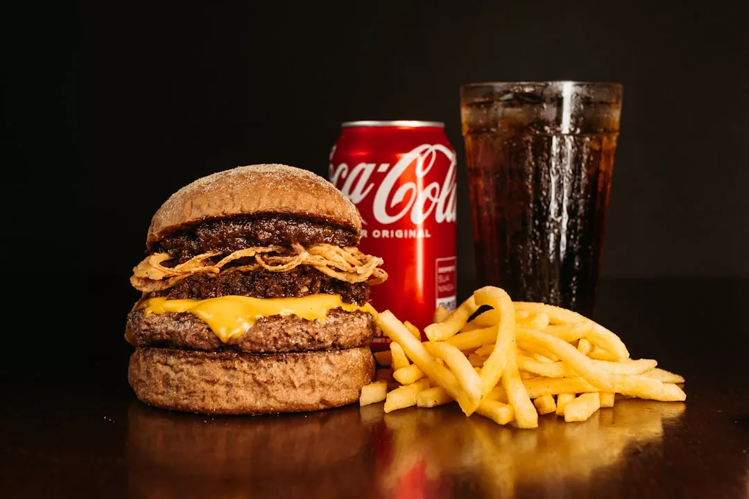 Ein Cheeseburger mit knusprigen Zwiebeln, eine Portion Pommes frites, eine Dose Coca-Cola und ein Glas Cola sind auf einer dunklen Fläche vor schwarzem Hintergrund angeordnet - perfekt für alle, die Tipps vor dem Start eines Abnehmprogramms suchen.