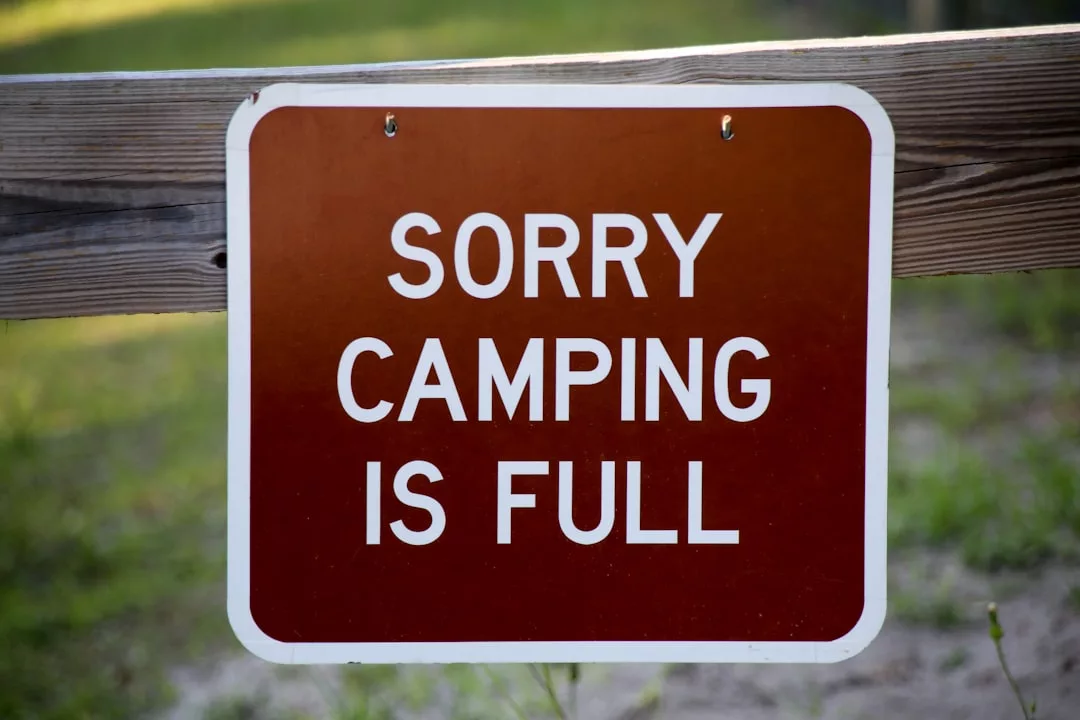 Ein braunes Schild mit weißer Schrift mit der Aufschrift "SORRY CAMPING IS FULL" ist an einem Holzzaun angebracht und erinnert Unternehmen an die Bedeutung einer effektiven Budgetplanung für eine bessere Ressourcenverteilung. Im Hintergrund ist Gras zu sehen.