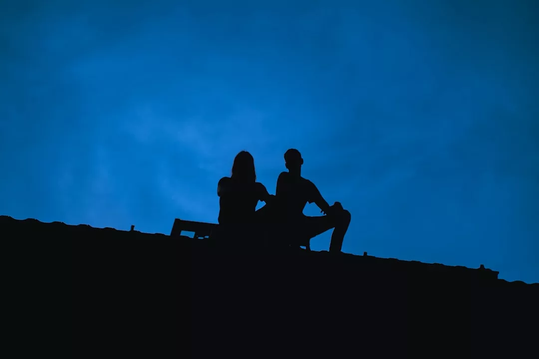 Zwei Menschen sitzen in der Abenddämmerung auf einem Dach, ihre Silhouetten zeichnen sich gegen einen tiefblauen Himmel ab - ein stiller Moment voller Nähe, der an eine glückliche Partnerschaft erinnert und wertvolle Tipps aus der Eheberatung erahnen lässt.