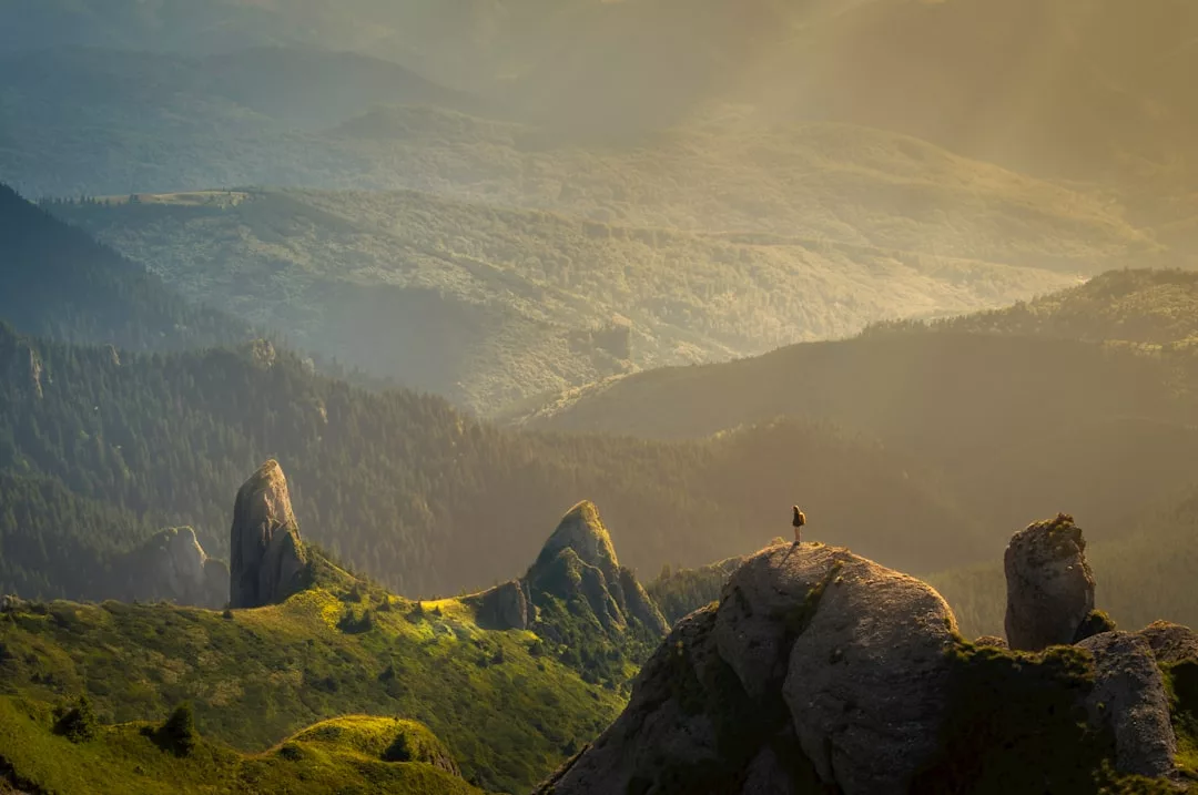 Eine Person steht auf einem Felsvorsprung mit Blick auf grüne Hügel, Wälder und Berge, die in warmes, goldenes Licht getaucht sind - eine inspirierende Kulisse für Fernarbeit und effektive Gestaltung.