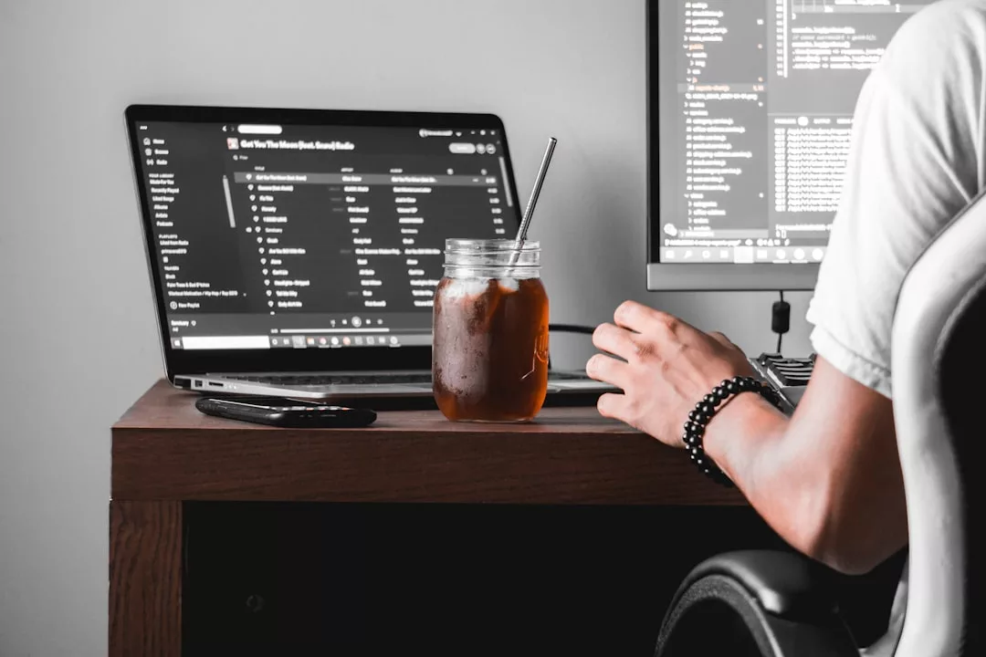 An einem Schreibtisch sitzende Person mit einem Laptop, einem Desktop-Monitor, auf dem ein Code angezeigt wird, der Produktivitätssteigerung durch effektive Methoden vorstellt, daneben ein Glas Eistee und eine TV-Fernbedienung auf einer Holzoberfläche.