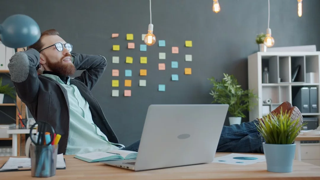Man sitzt entspannt am Schreibtisch, Füße hochgelegt, Hände hinter dem Kopf. Laptop und Papiere auf dem Tisch, Haftnotizen an der Wand - perfekte Umgebung für neue Strategien zur Mitarbeitermotivation im Unternehmen.