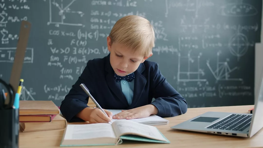 Ein Junge im Anzug schreibt in ein Notizbuch an einem Schreibtisch mit Büchern und einem Laptop und zeigt effektive Studierfähigkeiten vor einer Kreidetafel mit mathematischen Diagrammen und Gleichungen.