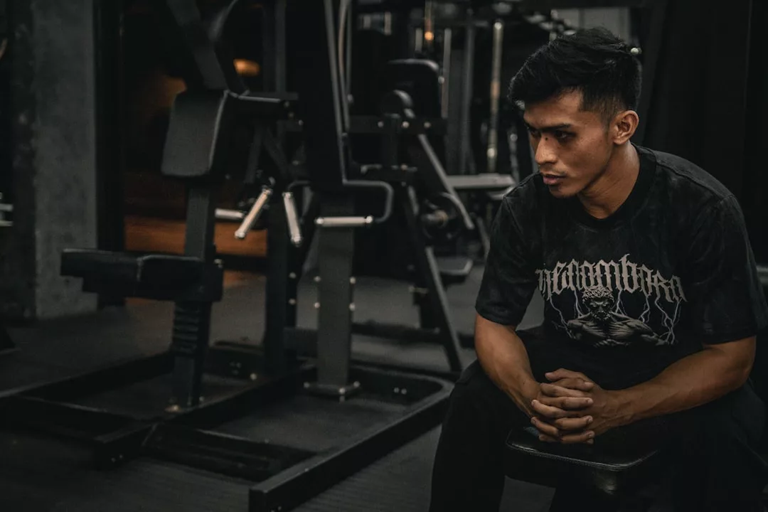 Ein Mann in einem dunklen grafischen T-Shirt sitzt auf einer Bank in einem Fitnessstudio und schaut mit ernster Miene nach unten. Im Hintergrund sind Fitnessgeräte zu sehen, die sich perfekt für effektive Workouts oder den Dreh von inspirierenden Workout-Videos eignen.