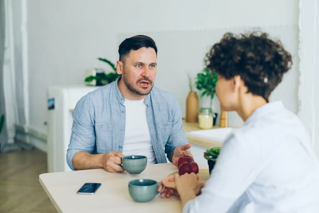 Zwei Personen sitzen an einem Tisch und unterhalten sich mit Kaffeebechern über Partnerschaft. Auf dem Tisch liegen ein Smartphone und ein Teller mit Obst. Im Hintergrund sind Küchenutensilien und Pflanzen zu sehen, die eine gemütliche Beziehungscoaching-Situation suggerieren.