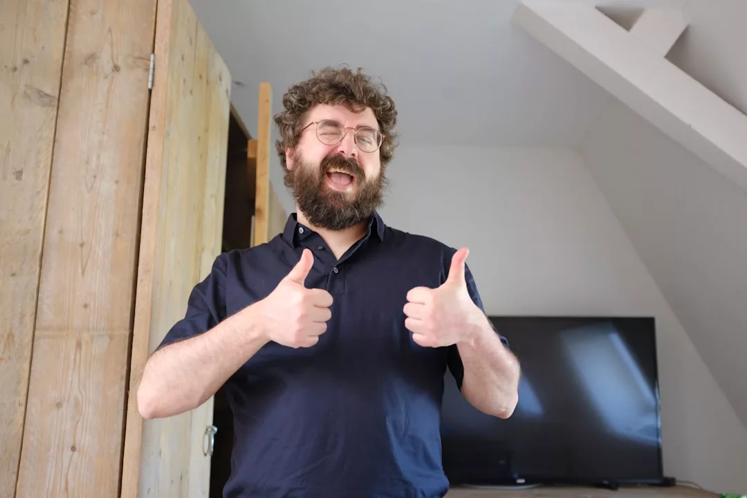 Ein Mann mit lockigem Haar und Bart steht in einem Haus, lächelt mit geschlossenen Augen und zeigt zwei Daumen nach oben - ein perfektes Bild für die gute Stimmung nach einem effektiven Feedback. Im Hintergrund sind ein Fernseher und eine Holztür zu sehen.