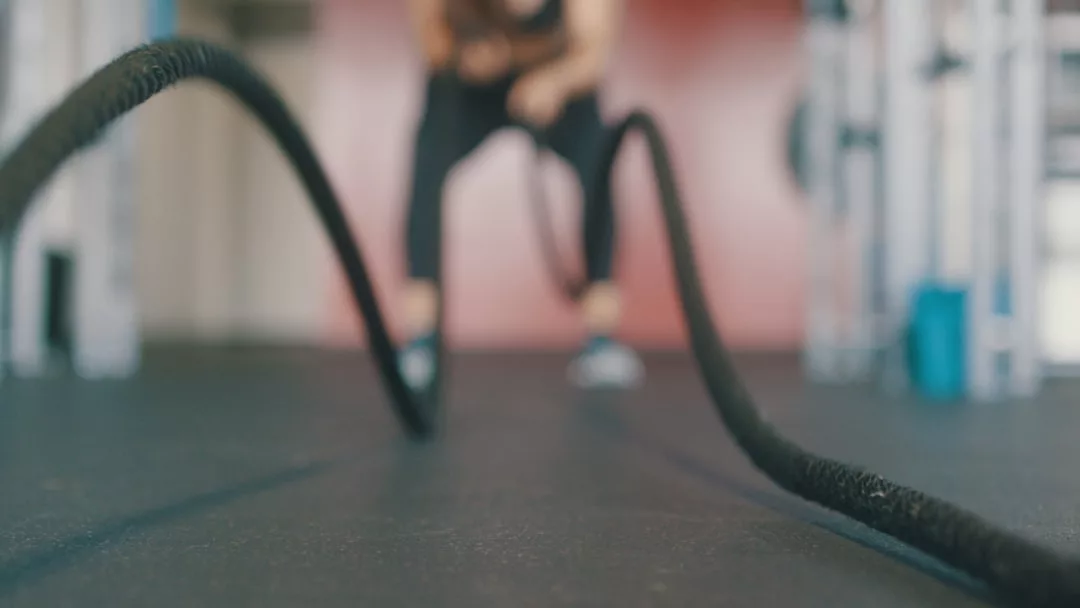 Eine Person demonstriert Fitness-Training mit Kampfseilen auf dem Boden einer Turnhalle, wobei die Seile im Mittelpunkt stehen, während die Person im Hintergrund verschwommen ist.