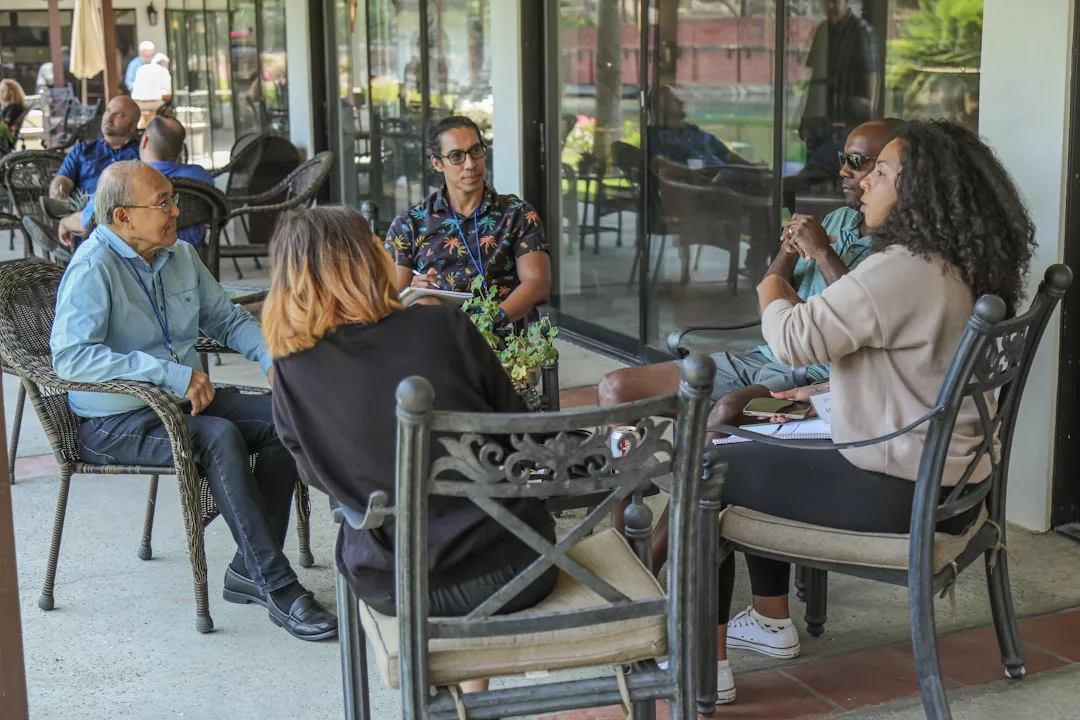 Fünf Personen sitzen im Kreis auf Terrassenstühlen vor einem Gebäude und unterhalten sich über Führungstraining, mit Notizbüchern und Papieren in der Hand.