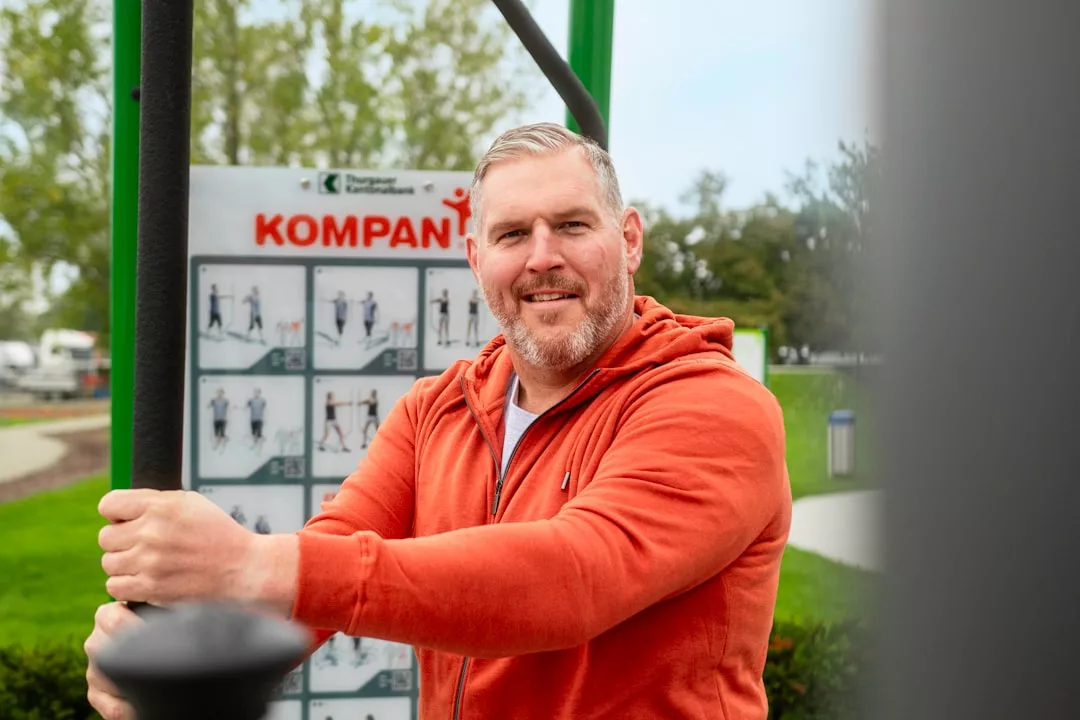 Ein Mann in einem orangefarbenen Kapuzenpulli hält sich an einem Outdoor-Fitnessgerät fest, während im Hintergrund ein Schild mit Tipps für aktives Training und grüne Bäume zu sehen sind.