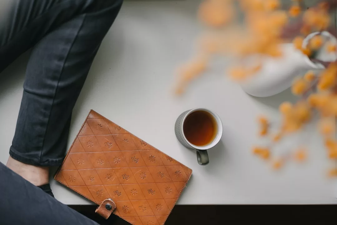 Ein braunes Ledernotizbuch, eine Tasse Tee und ein Teil der Beine einer Person in einer dunklen Hose liegen auf einem weißen Tisch - eine einladende Szene für das Selbststudium oder das Nachdenken über neue Methoden, mit einer verschwommenen Teekanne und orangefarbenen Blumen in der Ecke.