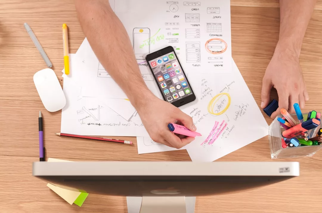 Person hebt Notizen zu App-Design-Skizzen hervor und nutzt dabei Tipps für besseres Zeitmanagement und gesteigerte Produktivität. Auf dem Schreibtisch liegen Smartphone, bunte Marker, Stifte, Maus und ein Computermonitor.