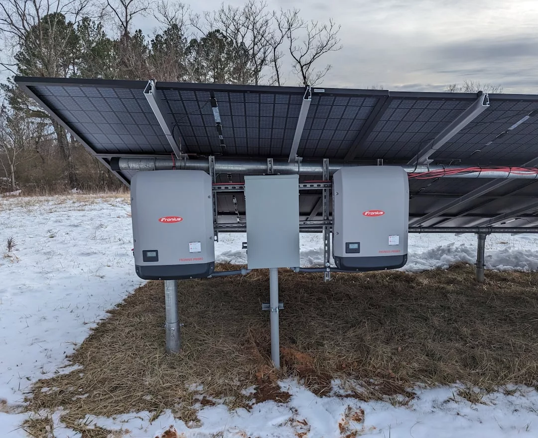 Zwei Solarwechselrichter, ein Schaltkasten und ein Energiespeicher sind unter Solarmodulen in einem verschneiten Außenbereich mit Bäumen im Hintergrund montiert.