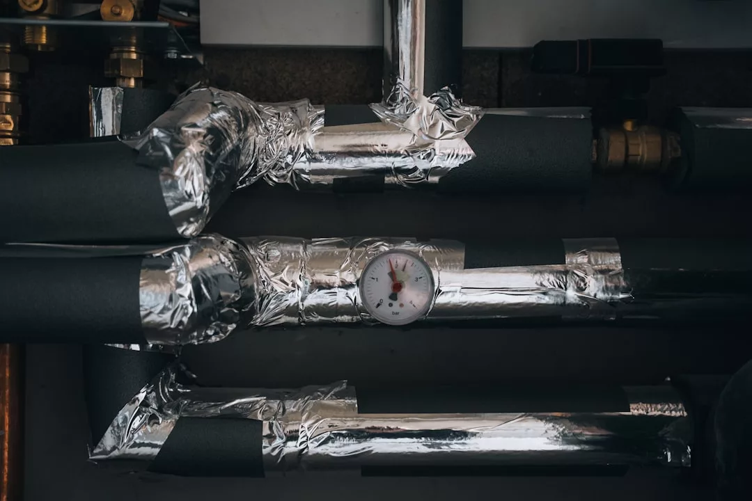 Drei isolierte Metallrohre, die mit reflektierendem Klebeband umwickelt sind, und ein rundes Manometer, das am mittleren Rohr befestigt ist, zeigen die Effizienz moderner Heizsysteme Zuhause in einer schwach beleuchteten Umgebung.