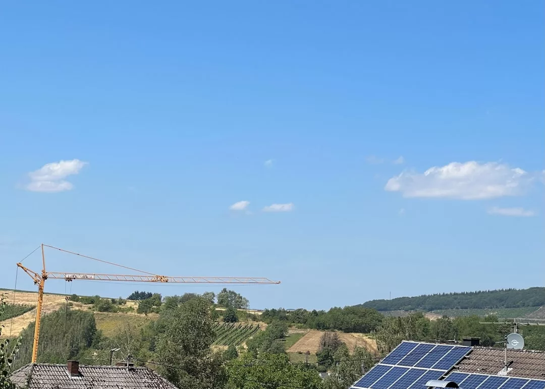 Eine Landschaft mit Dächern mit Solarmodulen und Solarstromspeichern im Vordergrund, einem Kran auf der linken Seite, Feldern, Bäumen und einem klaren blauen Himmel mit ein paar Wolken, die die effiziente Nutzung erneuerbarer Energien hervorheben.
