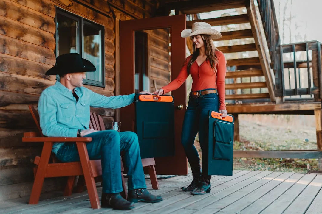 Ein Mann und eine Frau mit Cowboyhüten stehen auf einer hölzernen Veranda; der Mann sitzt, während die Frau ihm ein schwarzes, rechteckiges Objekt mit einem orangefarbenen Griff überreicht, das auf die Zukunft der Energiegewinnung mit effizienten Solarzellen hinweist.
