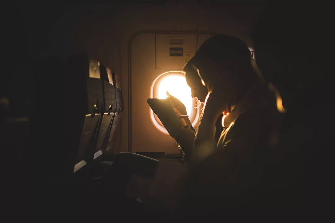Genieße deine perfekte Auszeit, während du am Fensterplatz eines Flugzeugs sitzt und ein Buch oder Gerät liest, beleuchtet vom Sonnenlicht, das durch das Fenster strömt.